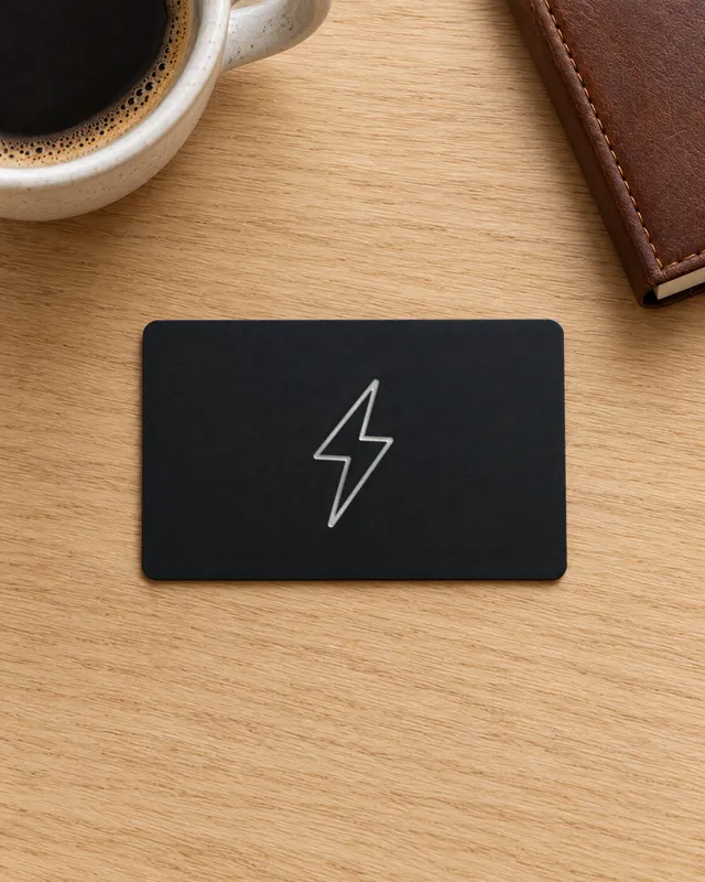 The matte black ZapCard with its silver foil bolt on an oak desk, seen from above, beside a coffee and a notebook